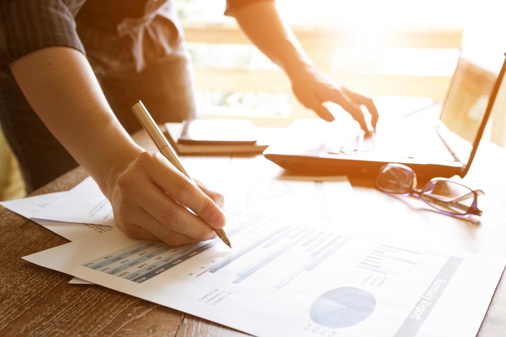 Frau mit Stift rechts über Papierunterlagen und am Laptop links bei der Arbeit